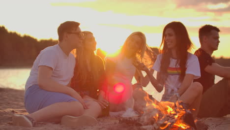 Two-men-and-three-young-women-are-sitting-around-bonfire-on-the-beach-with-beer.-One-of-the-women-is-telling-interesting-and-funny-story-at-sunset-in-summer-evening-on-the-lake-coast.